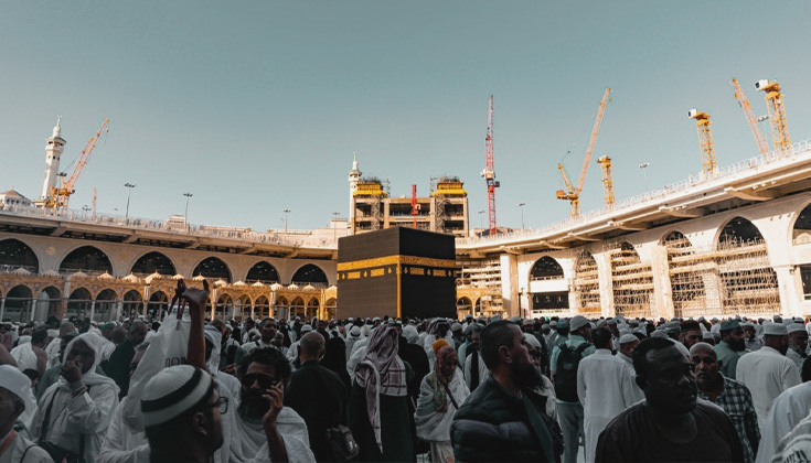 A large gathering of Muslim pilgrims near the Kaaba at Masjid al-Haram in Makkah, with construction cranes visible in the background.